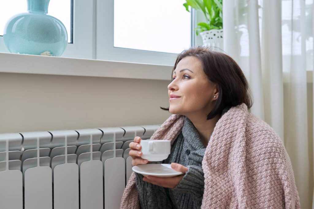 mujer abrigada dentro de casa tomando un café y mirando por la ventana